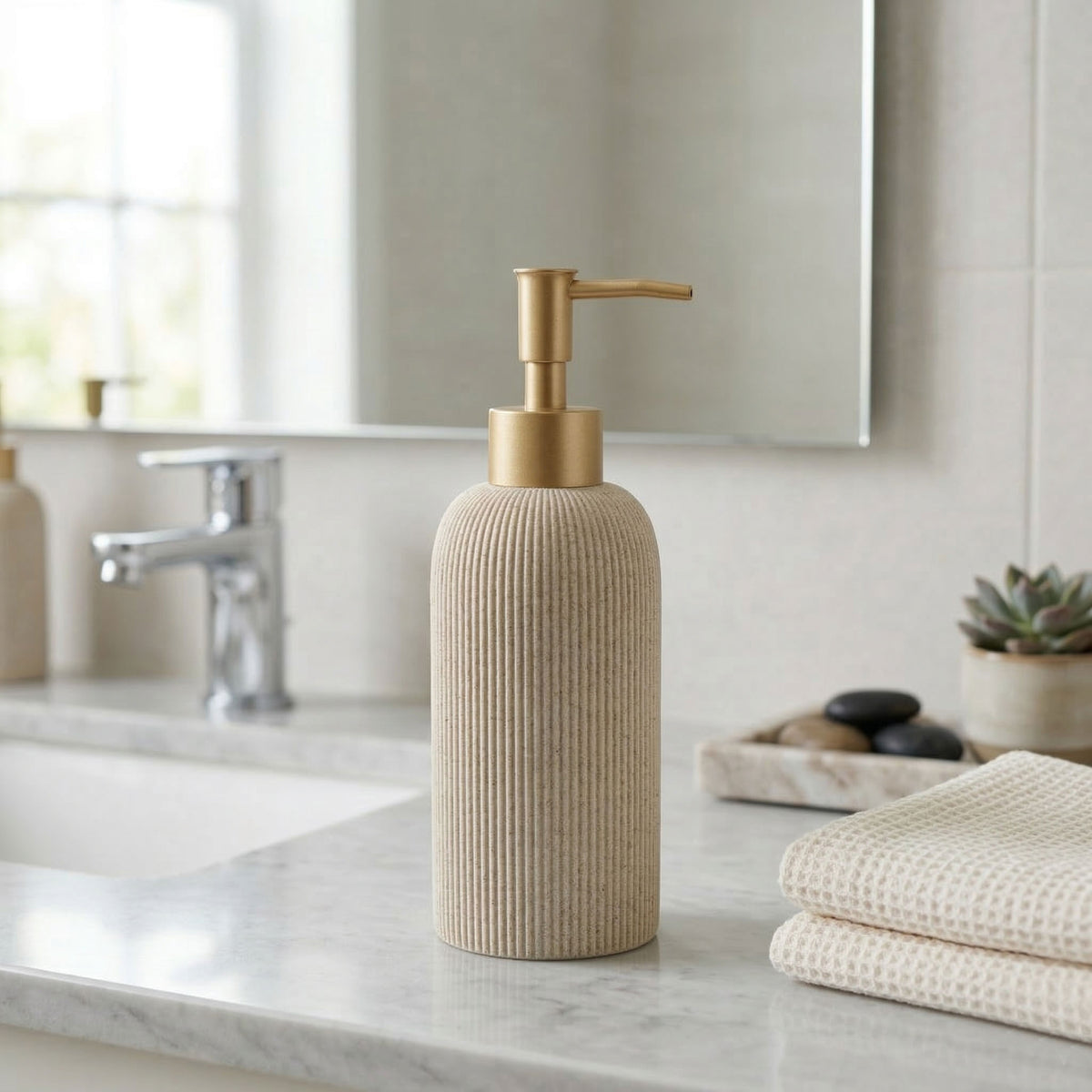 A ribbed beige soap dispenser with a mattegold pump, standing on a marble bathroom counter next to folded towels.