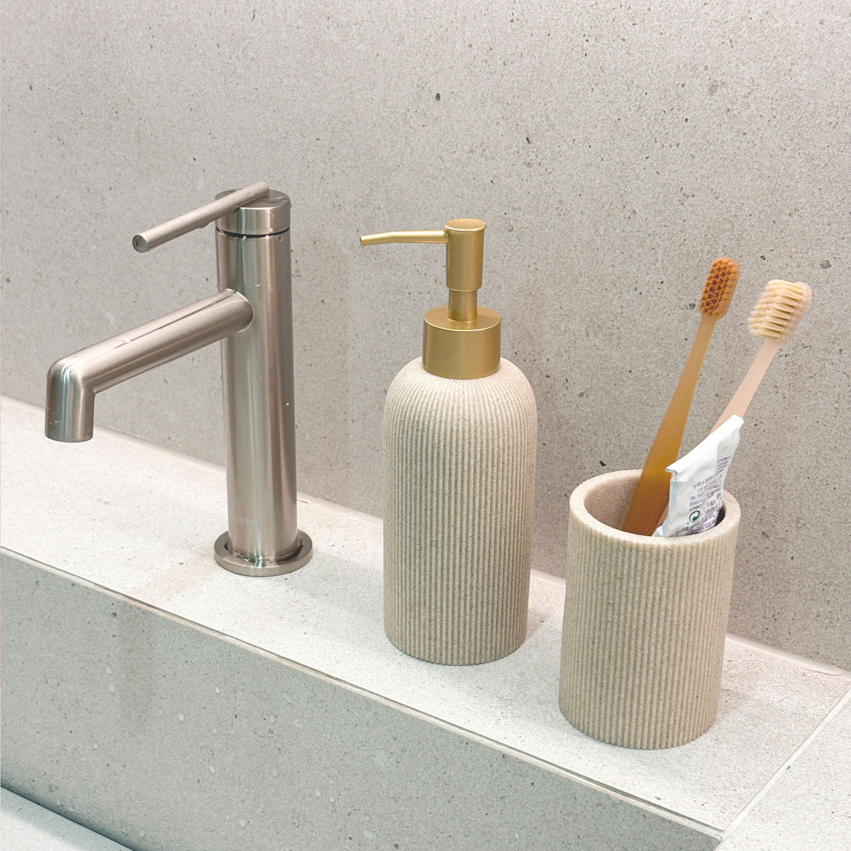Minimalistic bathroom sink setup with a faucet, sand beige soap dispenser with gold pump, and toothbrush holder.