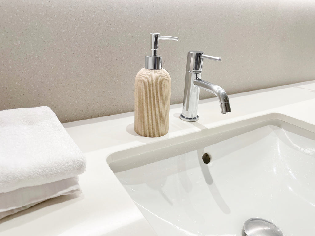 A ribbed textured beige dispenser bottle with a silver pump, styled on a white bathroom counter next to a chrome faucet and a folded white towel.
