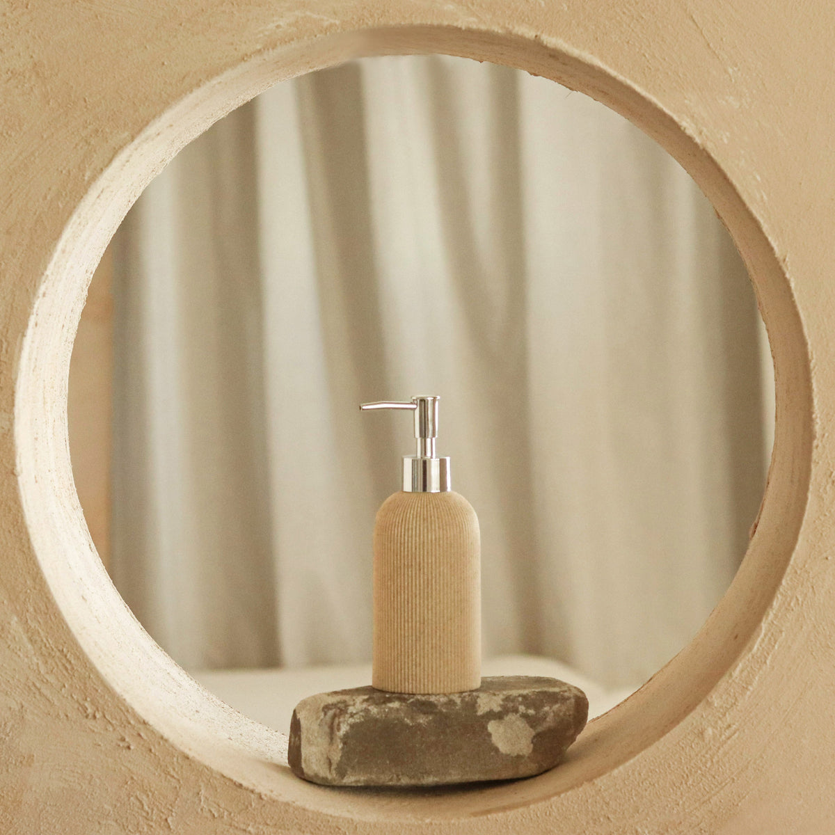 A beige ribbed pump bottle with a silver pump dispenser, resting on a small gray stone base inside a circular textured frame.